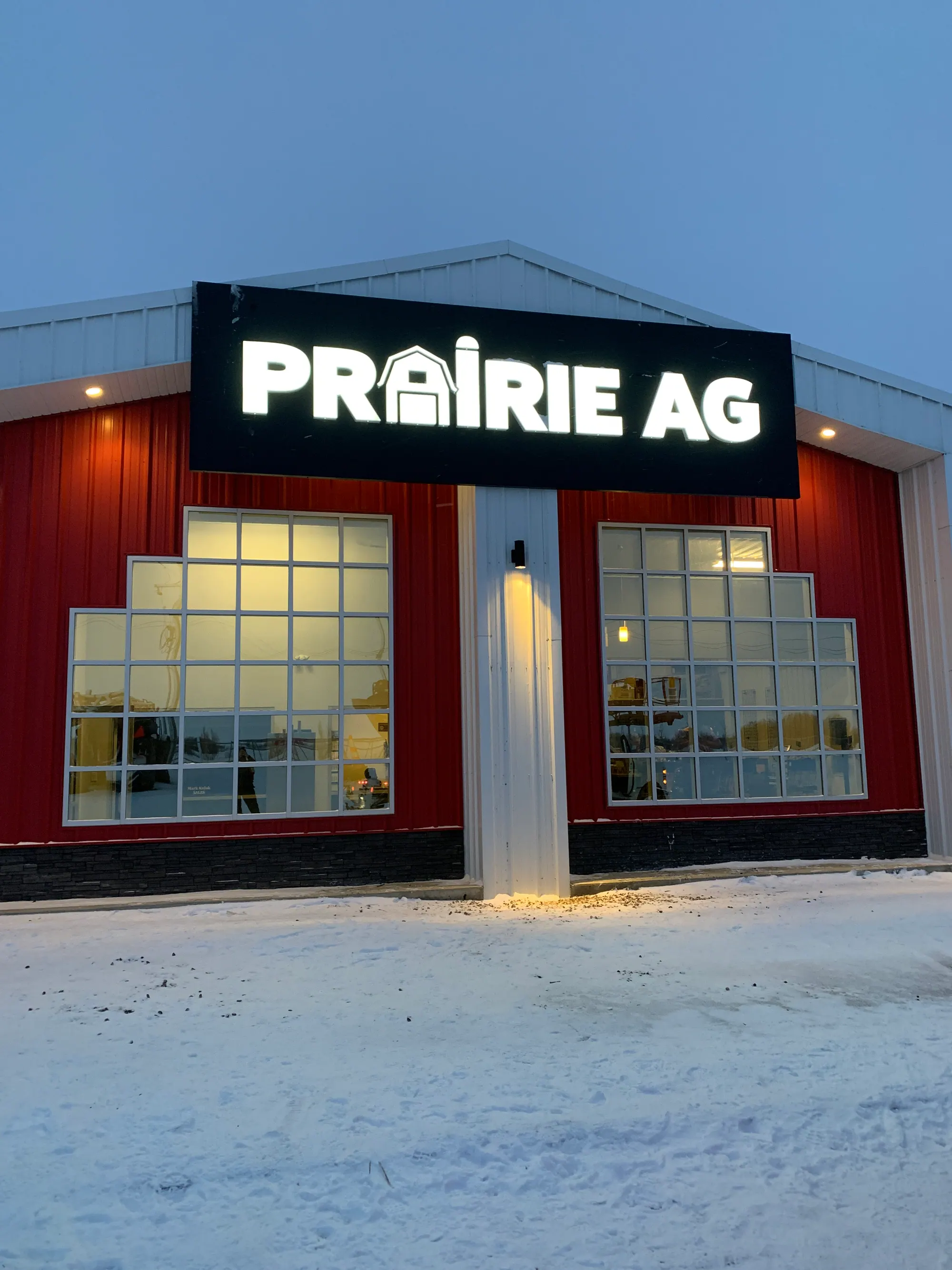 Prairie Ag dealership at dusk with the new illuminated sign above a red facade