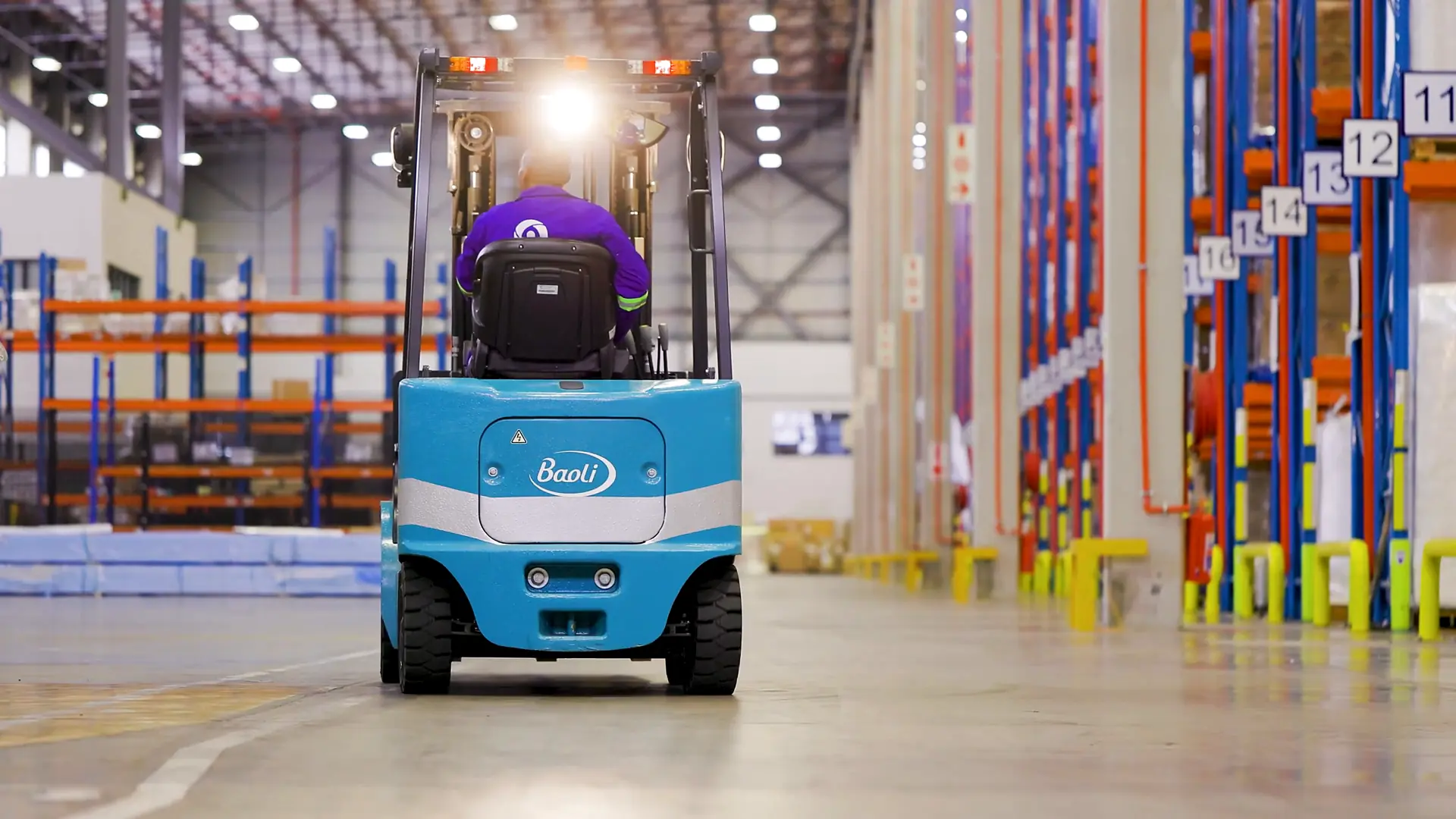 A teal Baoli electric forklift driving down a bright warehouse aisle, the Baoli logo on its counterweight