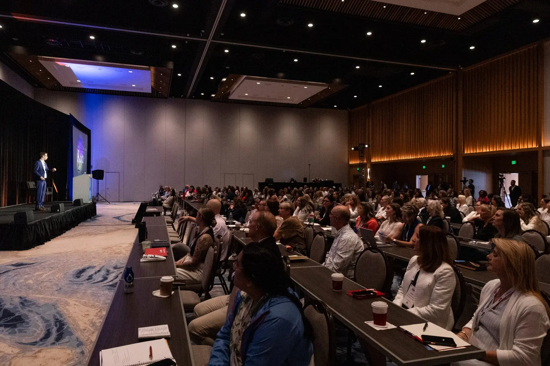 Michael Kutcher on stage in a navy suit, speaking to a full room under warm light