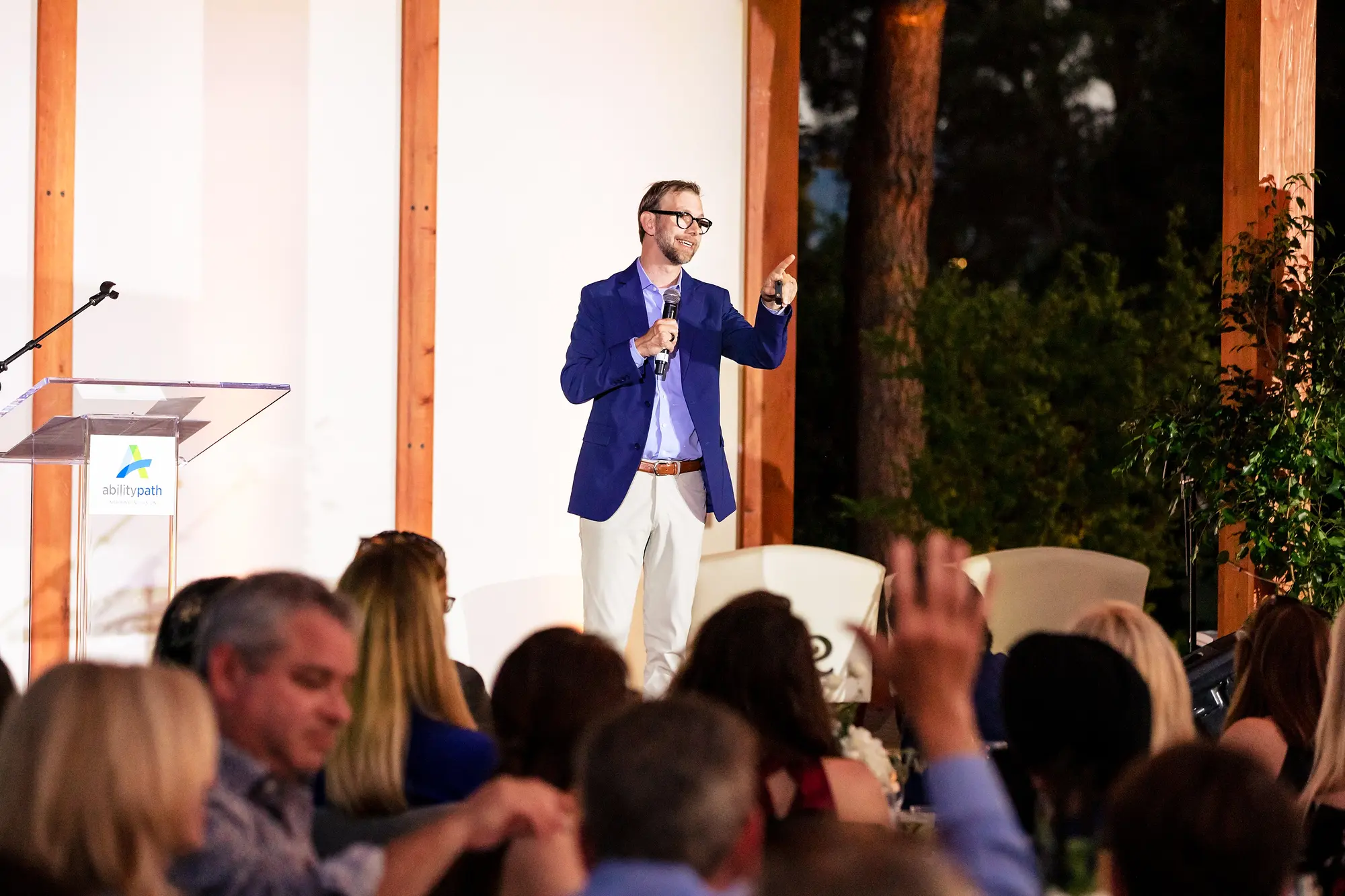 Michael Kutcher on stage with a microphone, an outdoor audience in the foreground with a hand raised